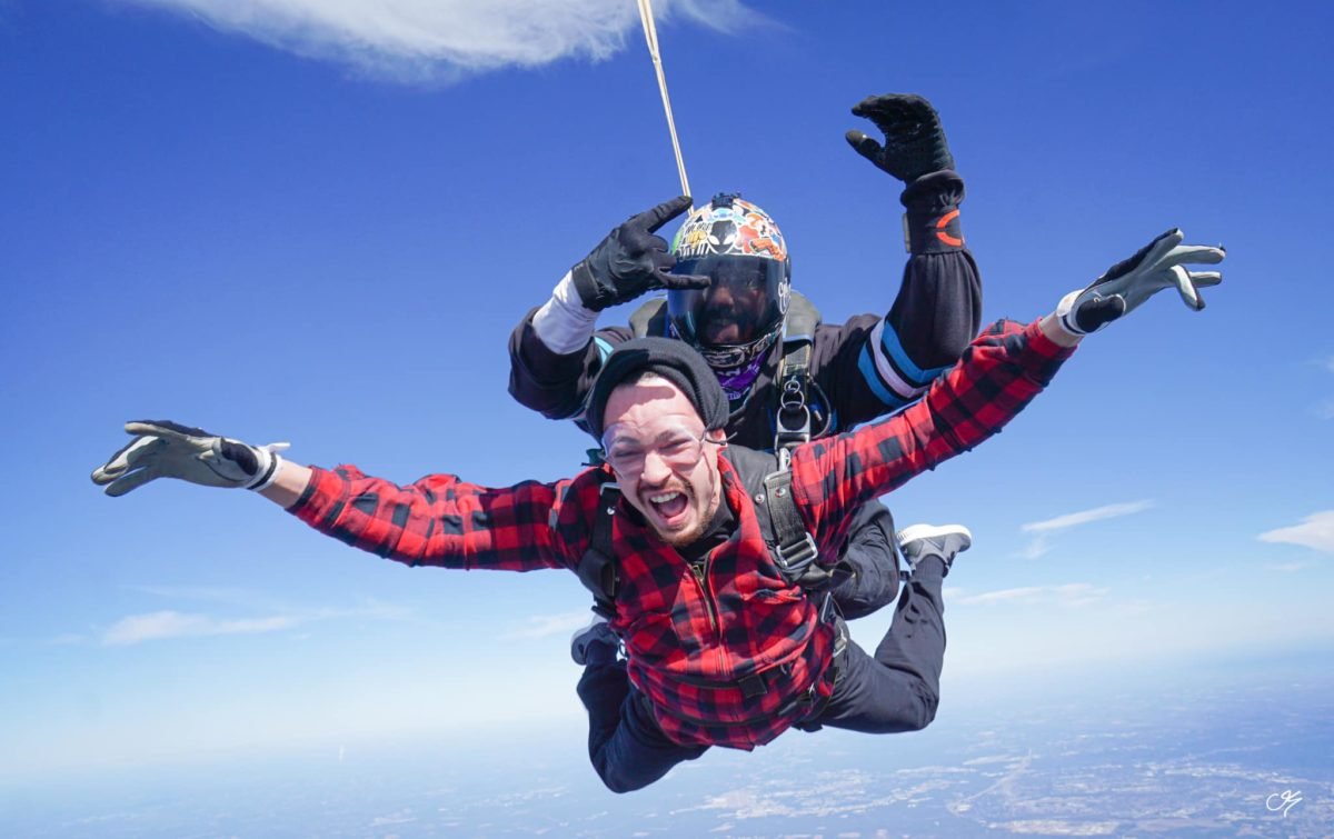 Male tandem student in red and black plaid shirt attached to a tandem instructor in freefall. 